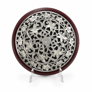 May include: A circular decorative plaque featuring a dark wood outer rim and a central silver-toned metal insert with an intricate openwork pattern of swirling vines and floral motifs set against a solid black background, displayed on a clear stand.