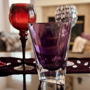May include: A deep purple glass vase with a thick clear base and a sticker reading LSA CRYSTAL VASE. The vase has a wide, angular opening and a smooth, reflective surface. It stands in front of a tall red glass goblet and a mosaic glass bowl.