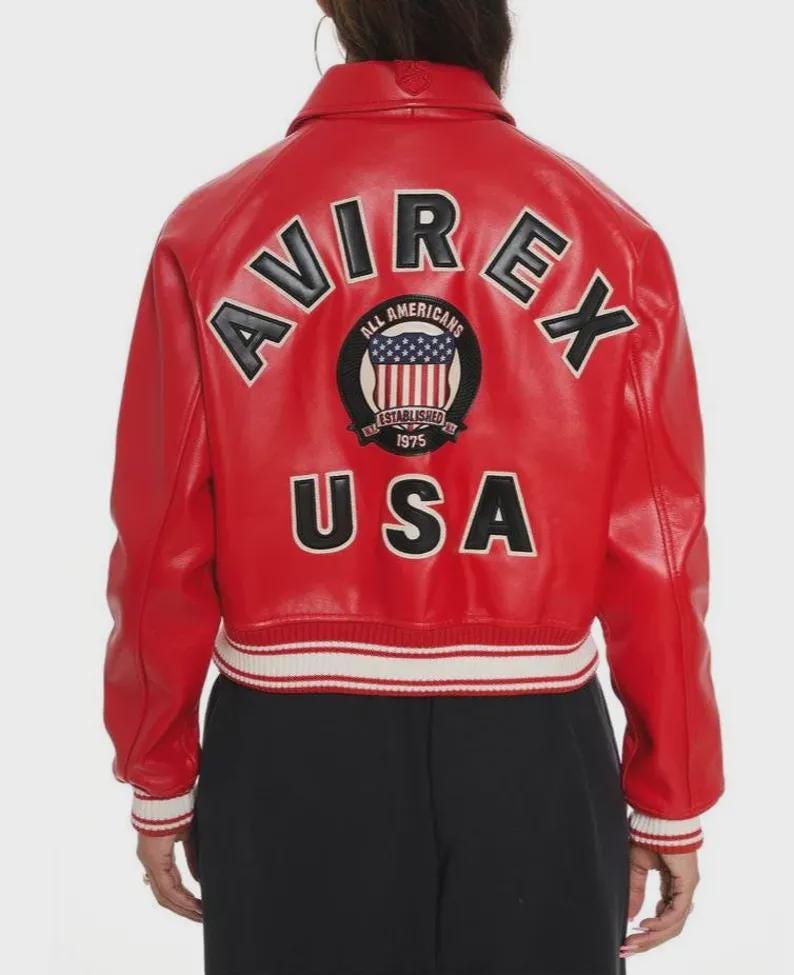 May include: A bright red leather jacket featuring the word AVIREX in large black letters across the upper back. Below this, a circular patch displays the American flag with the text ALL AMERICANS ESTABLISHED 1975. The word USA appears in black letters at the bottom, with white and red striped ribbed trim at the cuffs and waist.