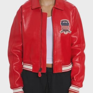 May include: A bright red leather jacket featuring a front zipper closure, a classic collar, and ribbed cuffs and hem with white stripes. On the left chest, there is a patch with the text Avirex, a shield graphic, and the words Established 1975.