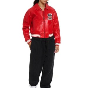 May include: A bright red leather bomber jacket with a front zipper, a collar, and striped ribbed cuffs and hem. The jacket features a patch on the left chest with the text "AVIREX" and a shield graphic. Paired with black sweatpants and white sneakers.
