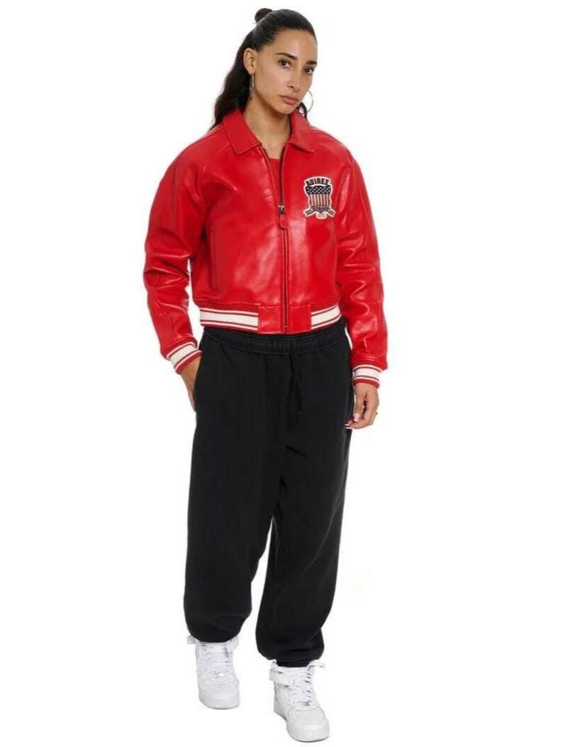 May include: A bright red leather bomber jacket with a front zipper, a collar, and striped ribbed cuffs and hem. The jacket features a patch on the left chest with the text "AVIREX" and a shield graphic. Paired with black sweatpants and white sneakers.