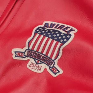 May include: A bright red leather jacket featuring an embroidered patch on the chest. The patch displays a shield with a blue field of white stars and red and white stripes, with the text AVIREX, ESTABLISHED 1975, and N.Y. in dark blue.
