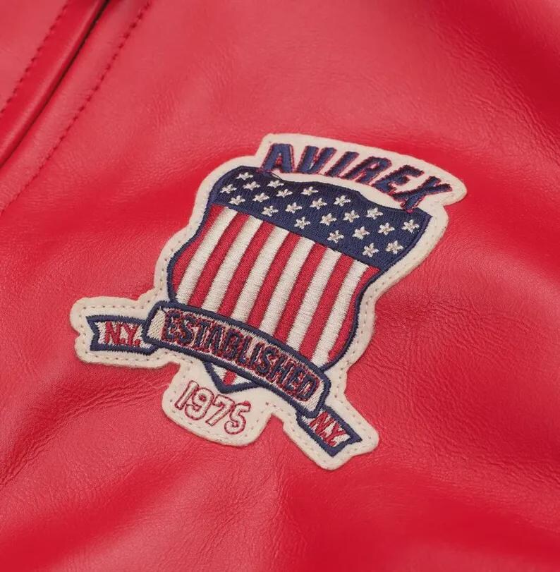 May include: A bright red leather jacket featuring an embroidered patch on the chest. The patch displays a shield with a blue field of white stars and red and white stripes, with the text AVIREX, ESTABLISHED 1975, and N.Y. in dark blue.