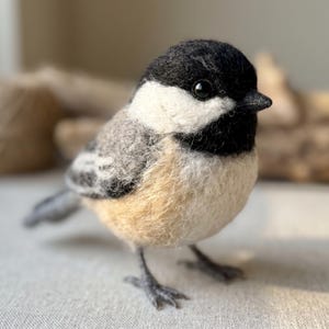 May include: A needle-felted chickadee figurine featuring a black cap and throat, white cheeks, and a grey back with tan-coloured underparts. The bird has a small black beak and dark, round eyes, standing on thin, dark grey textured legs.
