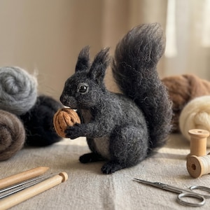 May include: A needle-felted squirrel figurine crafted from dark charcoal gray wool, featuring a bushy tail and pointed ears. The squirrel holds a small, light brown textured walnut in its front paws. The figure has a realistic appearance.