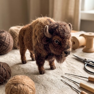 May include: A needle-felted brown bison figurine with a textured, woolly coat, small curved horns, and dark eyes. The bison stands on four legs, showcasing detailed craftsmanship with a dense, fuzzy appearance and a prominent hump on its back.