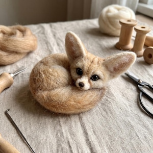 May include: A needle-felted fennec fox figurine with light tan and cream-colored wool fibers, large pointed ears, and dark eyes. The fox is curled into a circular shape with its tail wrapped around its body, showcasing a soft, textured appearance.