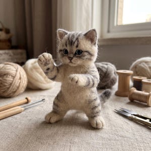 May include: A needle-felted kitten figurine with cream and gray tabby stripes, standing on its hind legs with one front paw raised. The kitten has dark eyes, a small nose, and a textured wool finish, capturing a realistic appearance.