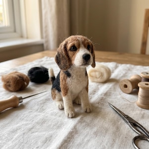 Puede incluir: Una figura de cachorro beagle de lana fieltrada con pelaje marrón, negro y blanco, ojos oscuros y nariz negra. El cachorro está rodeado de materiales de manualidades, incluyendo mechones de lana, carretes de madera, una aguja y tijeras.