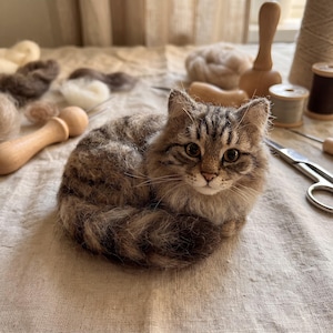 Puede incluir: Una figura de gato atigrado marrón y gris hecha con fieltro de aguja, con textura esponjosa, orejas puntiagudas y grandes ojos oscuros. El gato está en posición de descanso con la cola envuelta alrededor del cuerpo, mostrando pelaje detallado.