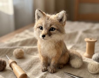 May include: A needle-felted fox figurine with textured tan, white, and grey wool fur, pointed ears, and dark eyes. The fox sits upright, surrounded by crafting supplies including a wooden handle needle tool, metal scissors, and small wool yarn balls.