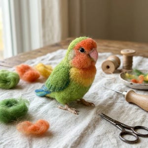May include: A needle-felted lovebird figurine with a bright green body, orange face, and yellow chest. The bird has a small pink beak and dark eyes. Nearby are loose tufts of green and orange wool, a wooden spool, a felting needle, and scissors.