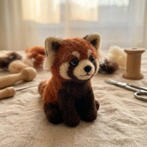 May include: A needle-felted red panda figurine featuring a textured coat of rust-orange, cream, and dark brown wool. The small animal has pointed ears, dark eyes, and a black nose, crafted with a soft, fuzzy appearance and a seated posture.