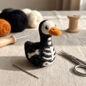 May include: A needle-felted duck figurine made of black wool with a white skeleton pattern on its chest and side. The duck has a white head, a bright orange beak, and two small black eyes. It is crafted from soft, fuzzy wool fibers.