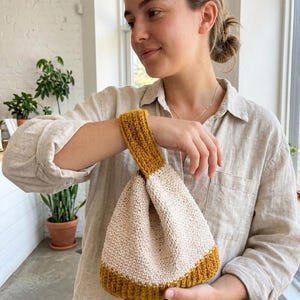 Peut inclure: Un petit sac crocheté à la main présentant un design bicolore avec un corps principal de couleur crème et une base ainsi qu'une anse de poignet jaune moutarde. Le sac est réalisé avec un point texturé dense et une poignée simple et fonctionnelle.