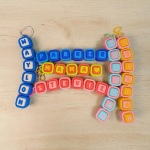 May include: A collection of colorful silicone bead keychains featuring square blocks with letters spelling out the names Waylon, Parker, Mama, Stevie, and Lillian. Each keychain is attached to a metal ball chain or a gold-toned lobster clasp.