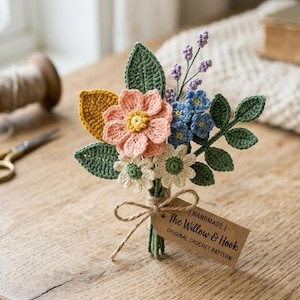 May include: A crochet flower bouquet featuring a large peach flower, small blue and white blossoms, and green leaves. The stems are tied with twine and a brown paper tag that reads "HANDMADE | The Willow & Hook | ORIGINAL CROCHET PATTERN".