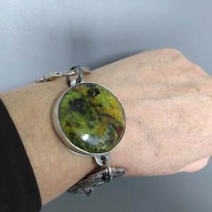 Op de afbeelding: Een armband met een grote, ronde, gepolijste steen in groen- en geeltinten, gezet in een zilverkleurige metalen rand. De armband bestaat uit gestructureerde, onregelmatige zilverkleurige metalen schakels die verbonden zijn door kleine ringen.