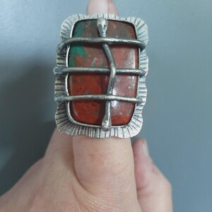 Op de afbeelding: Een rechthoekige ring met een grote, gladde, roodbruine en groene steen in een getextureerde, zilverkleurige metalen zetting. De steen wordt vastgehouden door een kooi-achtige constructie van dunne, gebogen, zilverkleurige metalen staven.