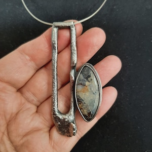 Op de afbeelding: Een hanger met een getextureerd, donker zilverkleurig metalen frame met een langwerpige rechthoekige opening. Aan de zijkant is een ovale steen bevestigd met gevlekte patronen in crème, bruin en zwart, gezet in een bijpassende metalen zetting.