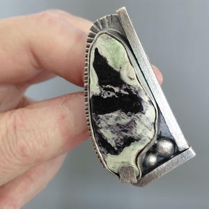 Op de afbeelding: Een opvallende ring met een grote, onregelmatige, gepolijste steen met gemarmerde patronen in lichtgroen, zwart en wit. De steen is gezet in een getextureerd, geoxideerd zilveren frame met een verticale balk, decoratieve inkepingen en twee zilveren bolletjes.