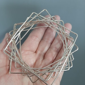 Op de afbeelding: Een set van meerdere in elkaar grijpende, vierkante armbanden gemaakt van gepolijst zilverkleurig metaal. De dunne, stijve vierkante frames zijn gelaagd en met elkaar verbonden om een sieraad te vormen met een gladde, reflecterende afwerking.