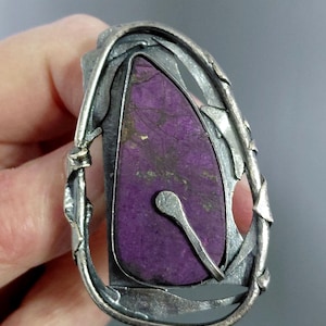Op de afbeelding: Een ring met een traanvormige paarse steen met donkere aders, gezet in een onregelmatig, getextureerd frame van geoxideerd zilver. Een dun, gebogen zilveren draadelement loopt over het oppervlak van de steen en zit vast in de zetting.