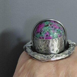 May include: A silver ring featuring a domed, polished ruby zoisite gemstone with vibrant magenta and green inclusions. The setting has a hammered, textured finish on the base and a smooth, arched silver wire frame surrounding the stone.