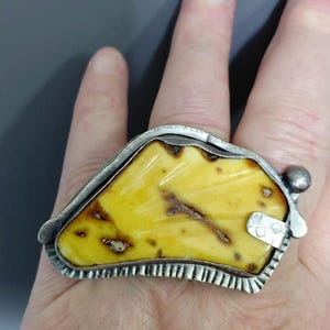 Op de afbeelding: Een grote statement ring met een onregelmatig, gepolijst stuk gele barnsteen met donkerbruine insluitingen, gezet in een getextureerd, geoxideerd zilveren frame met een gekartelde rand en een klein zilveren bolletje aan één kant.