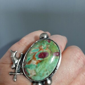 Op de afbeelding: Een ring met een grote, ovaalvormige groene en rode steen in een textuurrijke, donkerzilverkleurige metalen band. De metalen zetting bevat decoratieve bolvormige accenten en een klein, gestileerd dierenfiguurtje aan de zijkant van de steen.