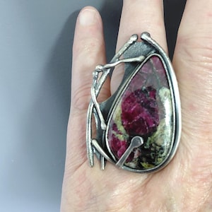 Op de afbeelding: Een grote statement ring met een traanvormige steen met diep magenta en donkergroene gevlekte patronen. De steen is gezet in een getextureerd, geoxideerd zilverkleurig metalen frame met organische, takachtige draadaccenten aan één zijde.