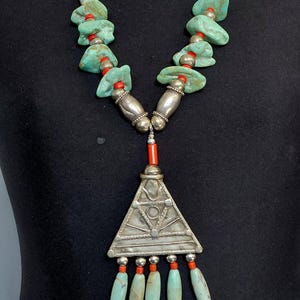 Op de afbeelding: Een ketting met turkoois stenen en kleine rode kralen aan een ketting. Het middelpunt is een grote, driehoekige zilverkleurige metalen hanger met gegraveerde geometrische patronen, geaccentueerd door vier bungelende turkoois kralen.