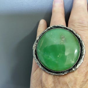 Op de afbeelding: Een grote ring met een gladde, ronde, levendig groene cabochonsteen in een textuurrijke, donker geoxideerde zilverkleurige metalen zetting. De zetting heeft een ruwe, onregelmatige rand die het gepolijste groene middendeel omringt.