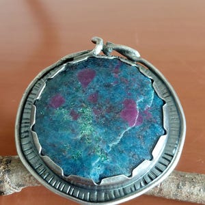 Pode incluir: Um pingente redondo com uma armação de metal prateado escuro texturizado e borda festonada. O centro contém uma pedra circular bruta com uma cor base azul-petróleo profunda, acentuada por inclusões irregulares de magenta e verde.