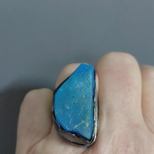 Puede incluir: Un anillo con una piedra grande de color azul brillante, de corte irregular y bruto, engastada en una banda de metal oscuro y texturizado. La piedra tiene una superficie mate y ligeramente porosa con variaciones de color, sujeta por un engaste.