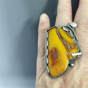 Op de afbeelding: Een grote, onregelmatige, gepolijste barnsteen gezet in een zilverkleurige metalen ring. De barnsteen heeft levendige gele en dieporanje tinten met natuurlijke insluitingen. De zilveren zetting heeft een getextureerde rand met twee kleine zilveren bolletjes.