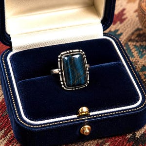 May include: A rectangular blue tiger's eye gemstone ring set in a silver-toned metal band with a beaded edge detail. The ring is displayed inside a dark blue velvet jewelry box with a white interior lining and a gold-toned metal clasp closure.