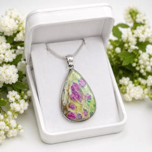 May include: A teardrop-shaped pendant necklace featuring a polished stone with vibrant magenta-pink inclusions set against a pale green and cream-colored matrix. The pendant is suspended from a thin silver-toned chain and rests inside a white box.