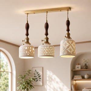 May include: A three-light pendant fixture featuring a rectangular brass ceiling plate. Each light hangs from a dark wood handle and brass socket, supporting a white ceramic shade with unique cut-out patterns: circular, grid, and floral designs.