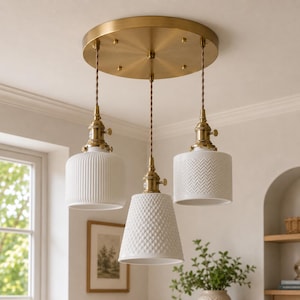 May include: A three-light pendant chandelier featuring a circular brushed brass ceiling canopy and three white ceramic shades with distinct textures. Each shade hangs from a twisted fabric cord connected to brass hardware and sockets.