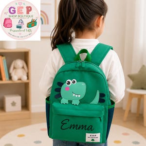 May include: A green backpack featuring a cartoon dinosaur graphic on the front pocket, with the name Emma printed in black script. The bag includes dark green side mesh pockets, adjustable shoulder straps, and a small white label with text.