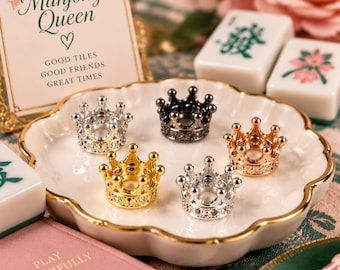 Peut inclure: Cinq petits supports en métal en forme de couronne aux finitions argentée, dorée, or rose et noire, ornés d'une bande de strass transparents. Ces objets décoratifs sont disposés sur une coupelle en céramique blanche à bordure dorée.