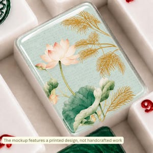 Puede incluir: Una ficha de mahjong rectangular con fondo verde menta claro y un dise&ntilde;o impreso de una flor de loto rosa, hojas verdes y follaje dorado similar a un helecho. La ficha tiene un acabado brillante e incluye texto sobre el dise&ntilde;o impreso.