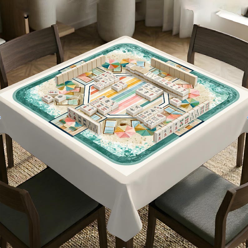 May include: A square table covered with a white tablecloth featuring a printed beach scene with sand, turquoise ocean waves, and colorful umbrellas. A set of white mahjong tiles with black and red characters is arranged on the tabletop surface.