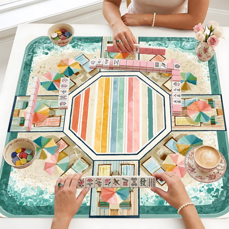May include: A square mahjong mat featuring a beach scene with colorful umbrellas and lounge chairs, printed with a central octagonal striped pattern. The mat is set with pink and white mahjong tiles, two bowls of colorful chips, and a cup of coffee.