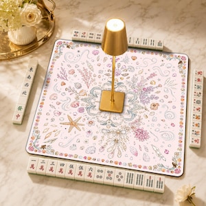 Peut inclure: Un tapis de mah-jong carré à fond rose clair orné d'une illustration détaillée de coquillages, de coraux et de motifs floraux dans des tons pastel. Une lampe dorée est placée au centre, entourée de quatre rangées de tuiles de mah-jong blanches.