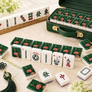 Puede incluir: Un juego de Mahjong con fichas blancas de reverso verde decoradas con flores de loto rosas y peces koi naranjas. El set incluye fichas con caracteres chinos en negro y rojo, una ficha de Joker y un malet&iacute;n verde oscuro con herrajes dorados.