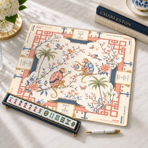 Op de afbeelding: Een vierkante mahjongmat met een crèmekleurige achtergrond en een chinoiserie-illustratie van apen in patronen, palmbomen en bloemmotieven. Een donkerblauw rekje bevat witte mahjongstenen met rode en groene karakters.