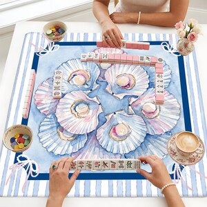 May include: A square mahjong mat featuring a watercolor print of seashells on a blue background with white and blue striped borders. The mat is set with mahjong tiles, two small bowls containing colorful game counters, and a floral teacup.